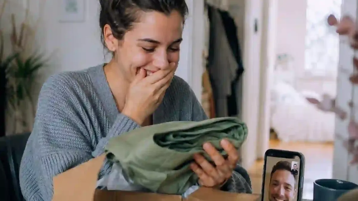 Eine Frau öffnet lächelnd ein Geschenkpaket, während sie mit ihrem Partner videotelefoniert