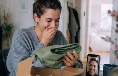Eine Frau öffnet lächelnd ein Geschenkpaket, während sie mit ihrem Partner videotelefoniert