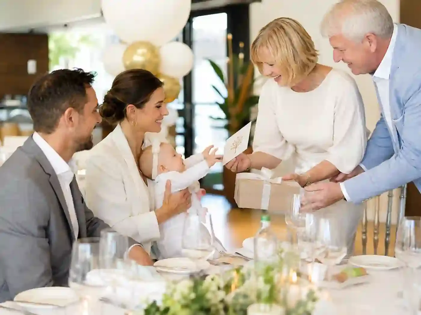 Familie übergibt lächelnd ein Geschenk mit Karte bei einer Tauffeier im Restaurant